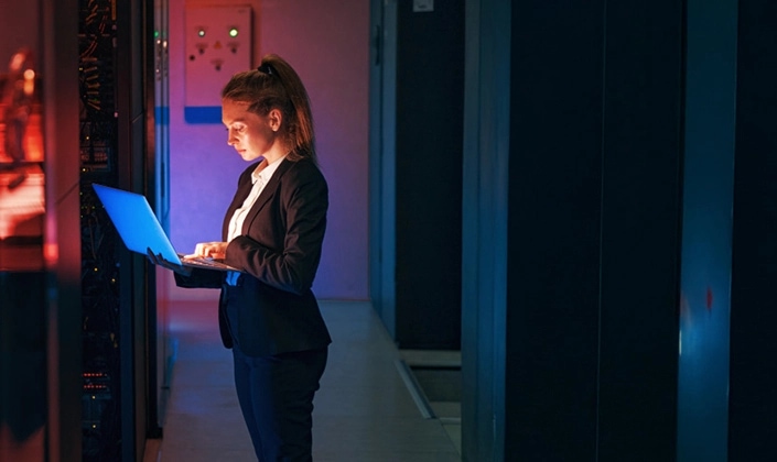 A woman in a professional suit working on a laptop in a dimly lit server room, surrounded by rows of illuminated server racks.