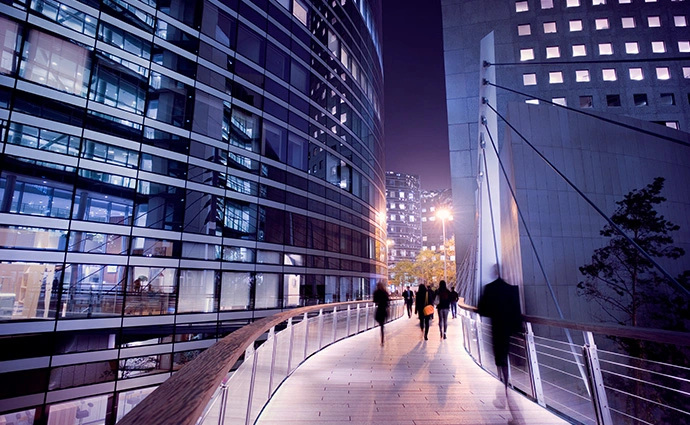 People walking on a bridge built between tall buildings lit with bright lights.