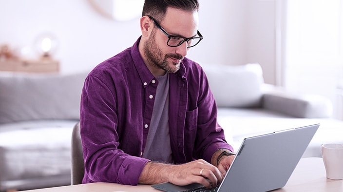 The image shows a man using a Lenovo laptop in a bright and modern environment. He is wearing a purple shirt and glasses, focusing on the screen while typing. In the background, a sofa and minimalist decor are visible, suggesting a comfortable and stylish workspace. Natural light enters through the window, creating a cozy atmosphere.
