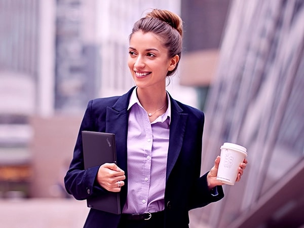 Close-up of a woman carrying coffee in her left hand and a Lenovo ThinkPad X13 Gen 4 laptop tucked under her right arm.