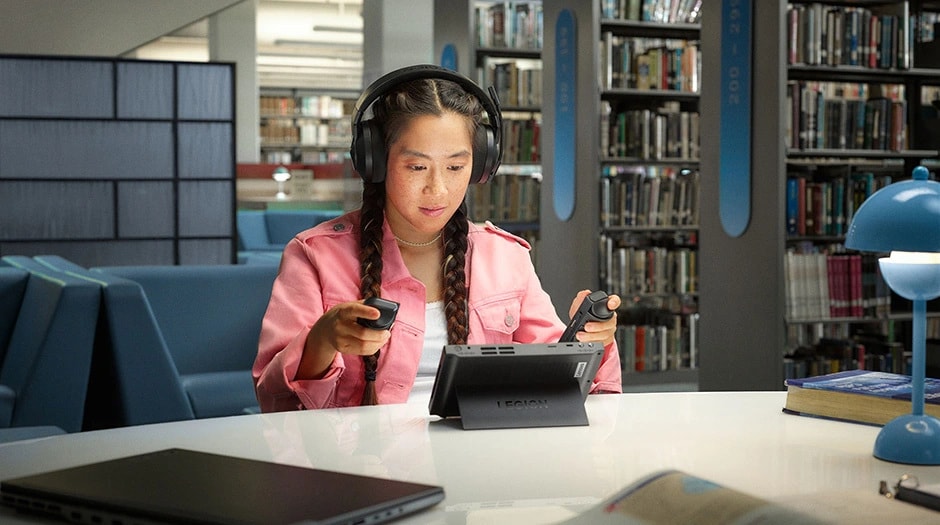 Person in library wearing headphones and playing on Legion Go handheld with detached controllers