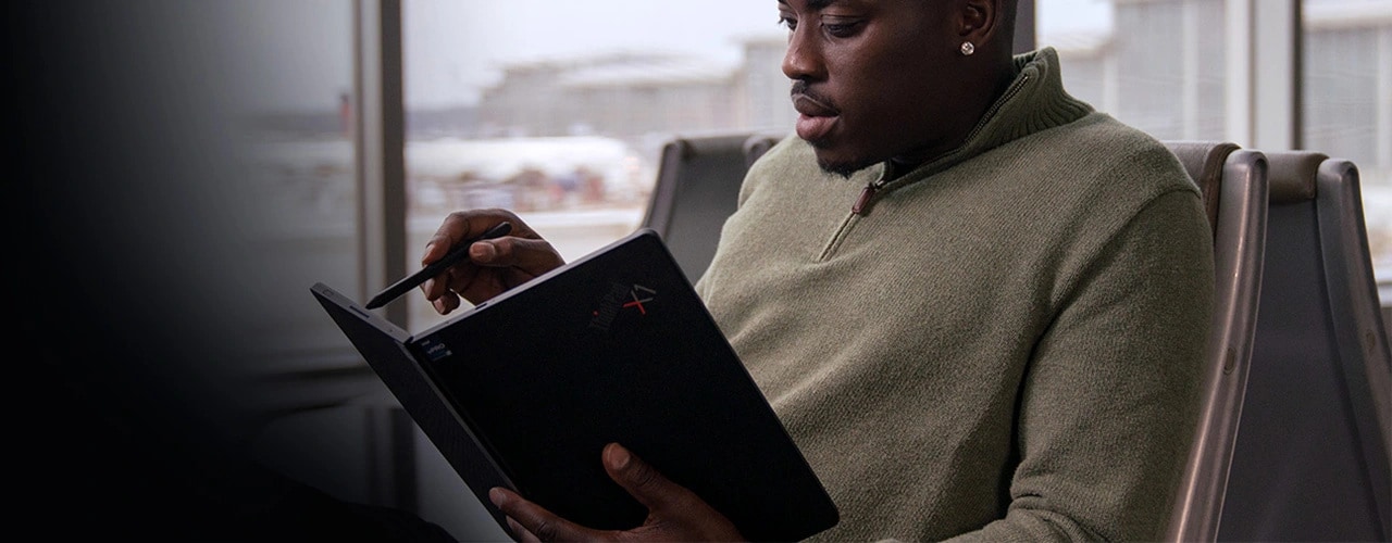 Uomo seduto su una sedia in aeroporto che usa il dispositivo Lenovo ThinkPad X1 Fold tenendolo aperto come un libro e scrive con una penna sul lato destro dello schermo.