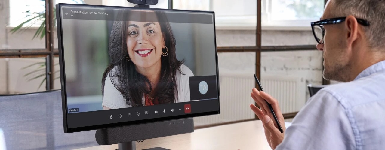 Lenovo ThinkSmart View Plus, with camera and soundbar attached, sitting on a desk in front of a gentleman communicating with a young woman via a virtual meeting software