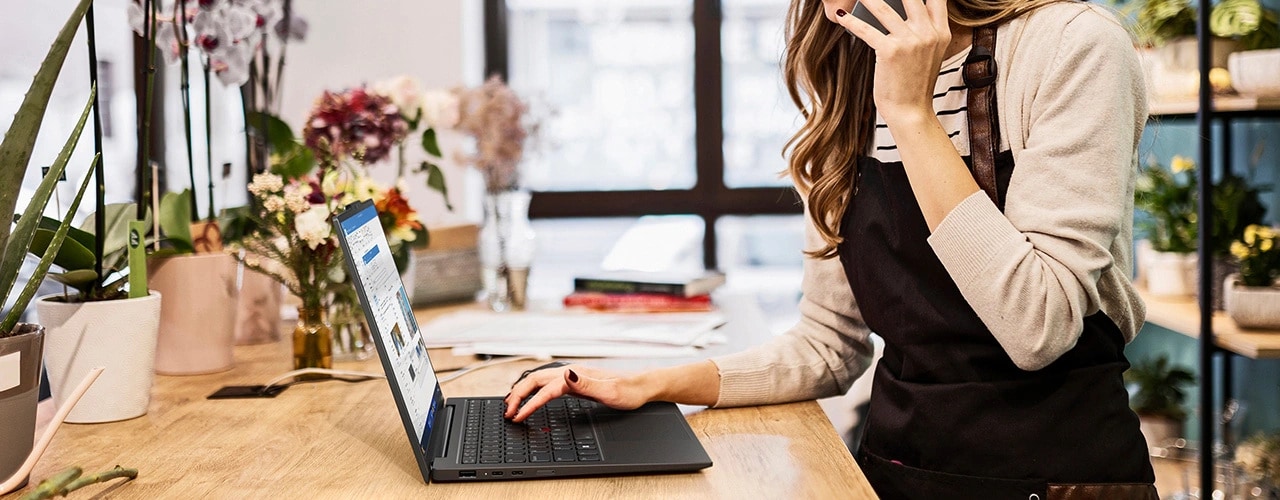 Kobieta korzysta ze smartfona i pisze prawą ręką na laptopie Lenovo ThinkPad X1 Carbon Gen 12, który stoi na stole otoczonym roślinami.