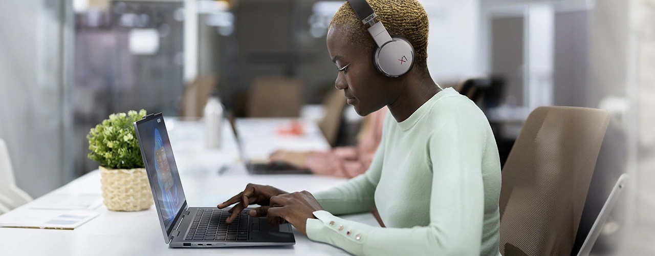 Eine Frau, die ThinkPad X1 Active Noise Cancellation Kopfhörer trägt, sitzt an einem Tisch und tippt auf dem wandelbaren Lenovo ThinkPad X1 2-in-1-Notebook, das von der linken Seite zu sehen ist.