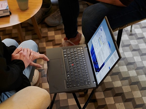 Overhead shot of right side profile of the Lenovo ThinkPad T14 Gen 5 (14 inch Intel) Eclipse Black laptop, placed in center & people sitting around it discussing work using Windows Copilot & Outlook email.