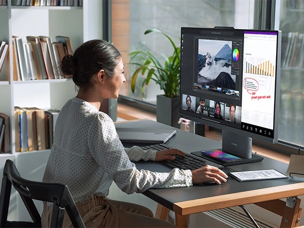 Lenovo ThinkCentre Neo 50a G5 27″ all-in-one desktop PC – front right view, showing a woman working on a software using wireless keyboard and mouse, along with having a video conference and the base wirelessly charging a mobile phone in her home office setup.