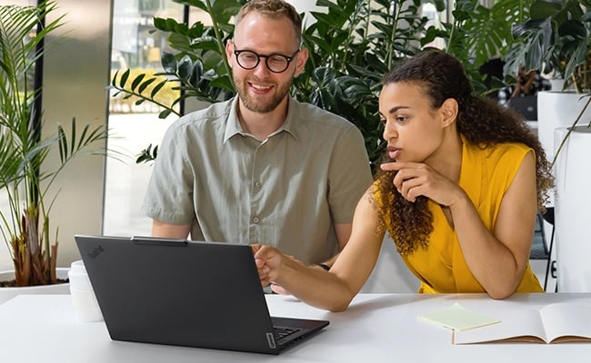 Two individuals discussing work on the Lenovo ThinkPad T14 Gen 5 (14” AMD) Eclipse Black laptop, focusing rear profile & its effective WiFi connectivity.