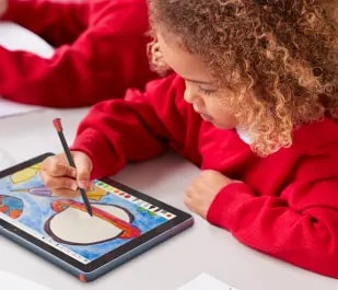 A young child in elementary school is seated at a table, using a stylus to draw on a tablet.