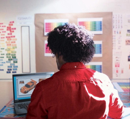 A designer working on a car model on his laptop with colorful notes pinned to a bulletin board in the background.