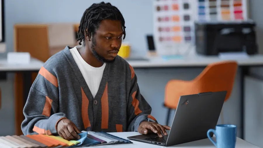 A man sitting at a table, looking at color swatches as he uses a Lenovo ThinkPad X1 laptop.