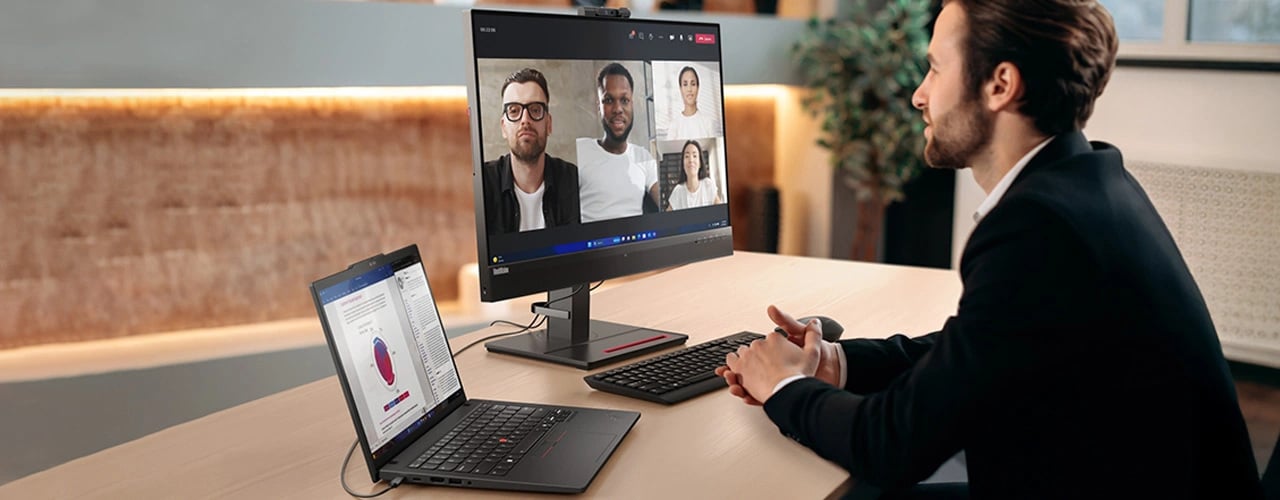 A person sitting at a desk, participating in a videoconference on a monitor connected to a ThinkPad T14 Gen 6 (14" AMD) laptop