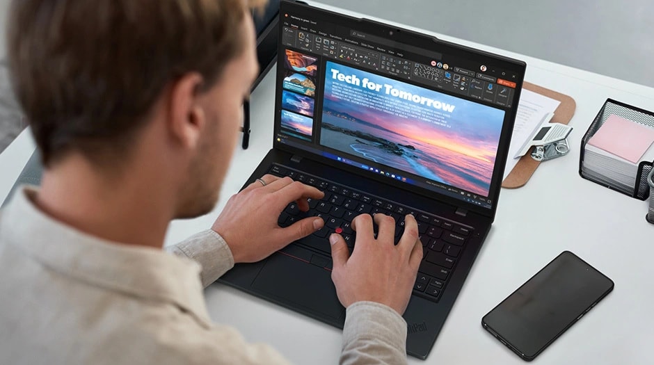 Lenovo ThinkPad L14 Gen 6 (14" AMD) laptop on a desk, seen over the shoulder of a person with both hands on the keyboard and a presentation deck open on the display