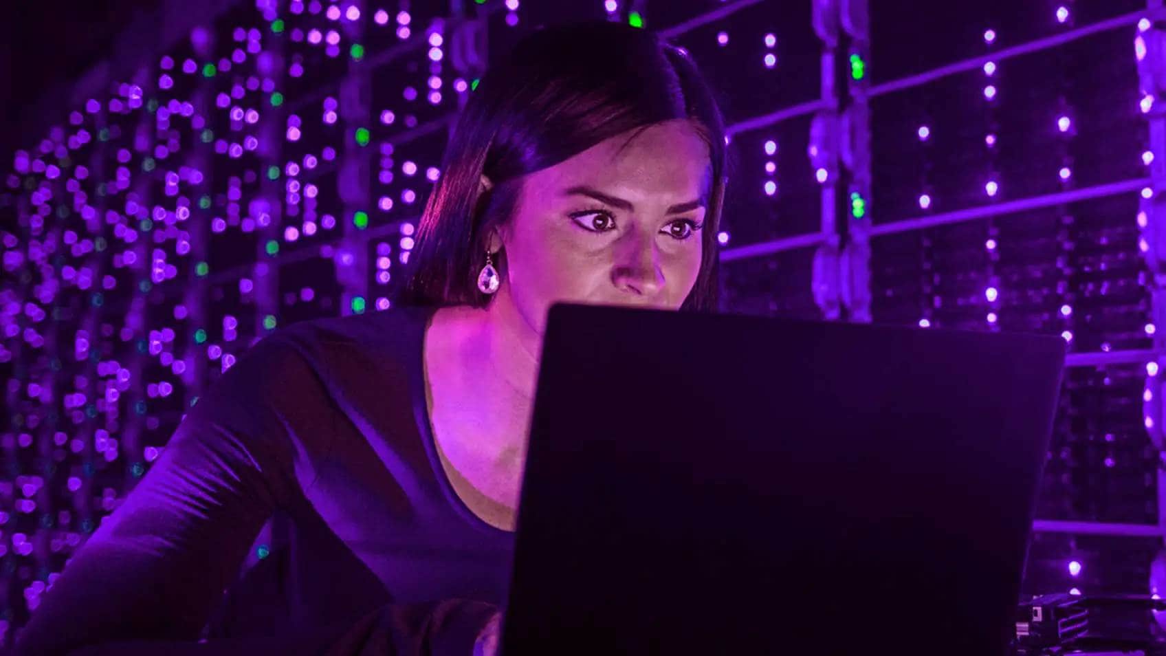 Une femme regardant attentivement l’écran de son portable avec des lumières pourpres des serveurs derrière elle.