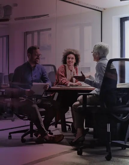 Trois collègues assis à une table dans un bureau moderne, souriant et discutant de leur travail.