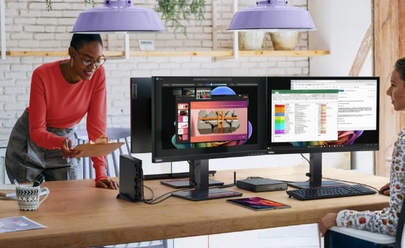 Two women across from each other at a table with three computer monitors and a Lenovo ThinkCentre neo desktop PC.