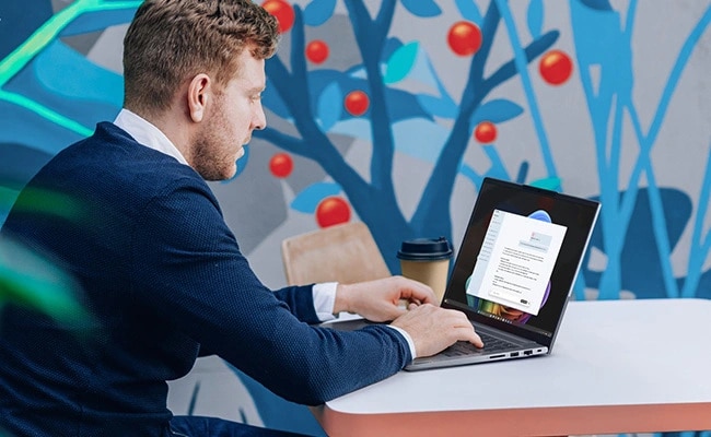 Ein Mann sitzt an einem Tisch und nutzt das Lenovo ThinkBook 14 Gen 9 Notebook, neben einem farbenfrohen Wandbild im Hintergrund.