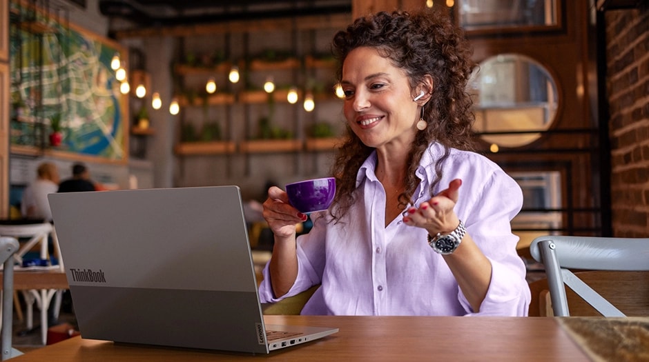 A woman in a café with earbuds faces forward gesturing while on a video call on  a Lenovo ThinkBook 16 Gen 9 laptop.	