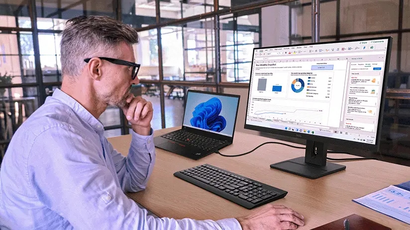 Hombre trabajando en un escritorio, viendo gráficos de datos y gráficos en un monitor grande, con un portátil y el teclado delante en un entorno de oficina moderno.