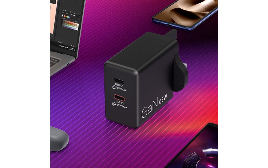 Black 65W GaN charger with dual USB-C ports, surrounded by a laptop, smartphones, and a mouse, on a purple-pink striped gradient background.