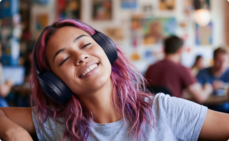 A woman with pink hair wearing black over-ear headphones smiles with her eyes closed in a café.