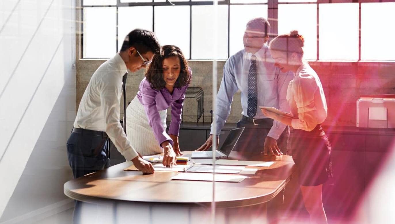 A diverse group of professionals collaborates around a conference table, reviewing documents in a modern, sunlit office.