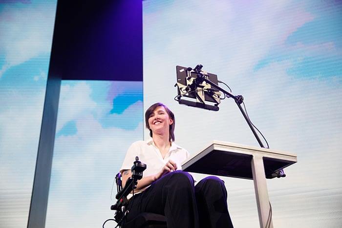 Erin Taylor sorrindo, em uma cadeira de rodas, usando uma tecnologia no palco durante uma apresentação.