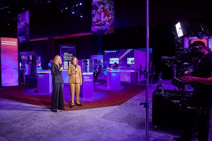 Laura Quatela and Mary Jacques on camera in a tech demo area, surrounded by purple lighting and AI infrastructure displays.