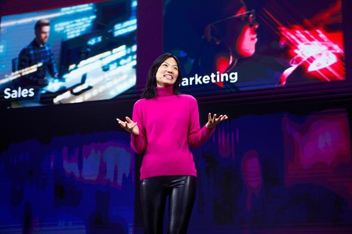 Linda Yao presenting on stage with a digital screen showing sales and marketing visuals behind her.