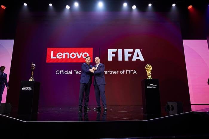 O CEO da Lenovo, Yuanqing Yang, e o presidente da FIFA, Gianni Infantino, apertando as mãos no palco, celebrando a parceria da Lenovo com a FIFA, com troféus em exibição.