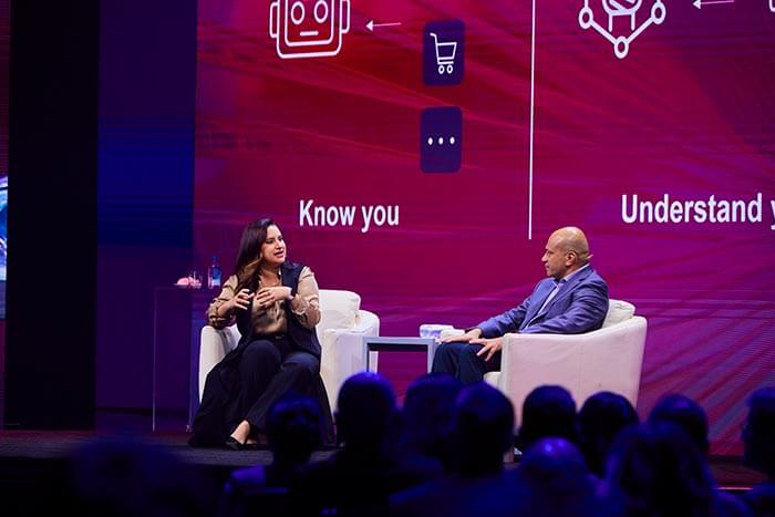 A CEO da Open Machine, Allie K. Miller, e o CTO da Lenovo, Tolga Kurtoglu, no palco discutindo IA em frente a um fundo com visuais de chatbot e carrinho de compras.