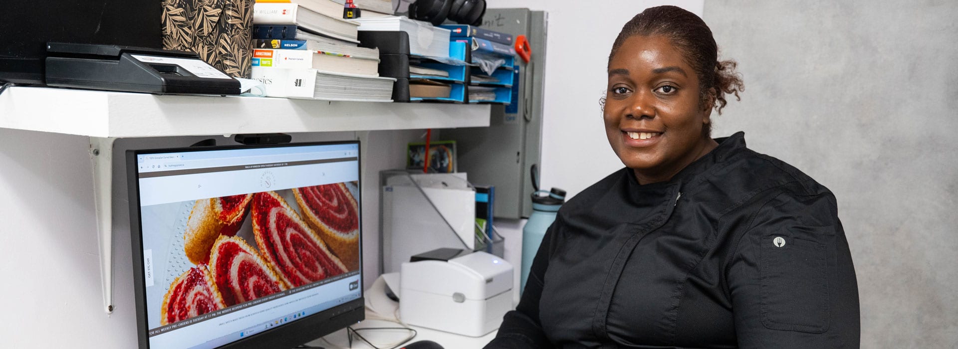 An African American woman wearing a black chef's jacket sitting in her business's office smiling at the camera.