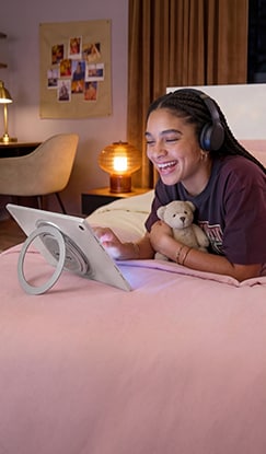 A woman lies in bed wearing headphones, watching a TV series on a Lenovo tablet.