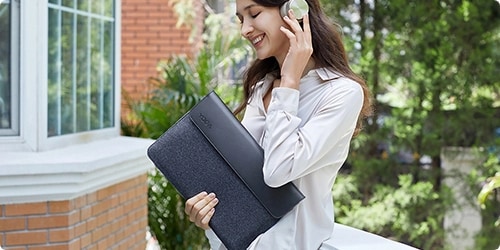 Woman in the street listening to music on the Yoga Active Noise Cancellation Headphones and holding a Yoga 14-inch Sleeve in her hand.