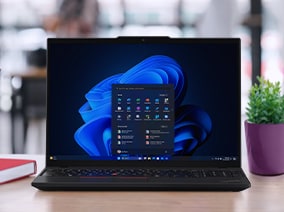 A black laptop on a wooden desk displaying the Windows 11 start menu with a blue abstract background, surrounded by office items including a pencil holder, stacked notebooks, and a small green plant in a purple pot.