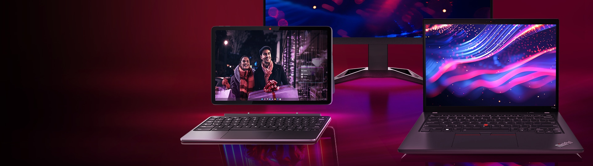 A Lenovo tablet with a keyboard and a Lenovo laptop are displayed against a deep burgundy background. The tablet shows an image of two people, while the laptop features vibrant, colorful graphics on its screen.