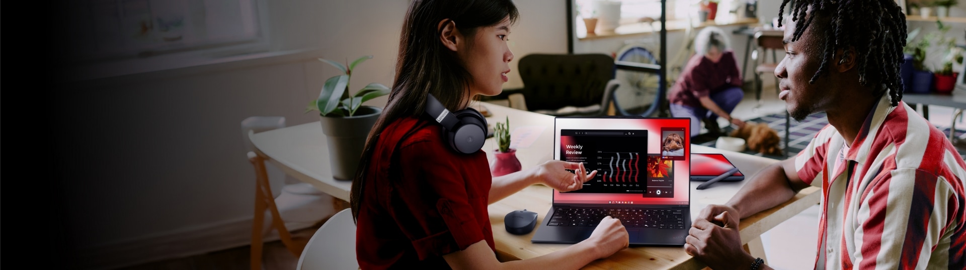 Une femme avec des écouteurs Lenovo, assis à une table, pointant vers un écran de portable ThinkPad, engagée dans une conversation avec un homme, dans une pièce avec d’autres personnes et un chien en arrière-plan pendant la journée.