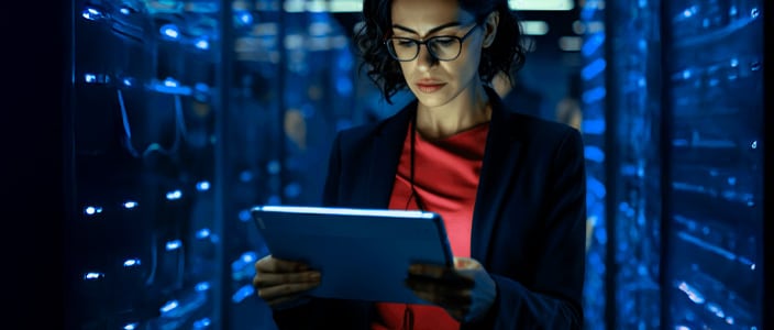 Person standing in a server room with blue-lit racks, holding a tablet while surrounded by rows of network equipment.