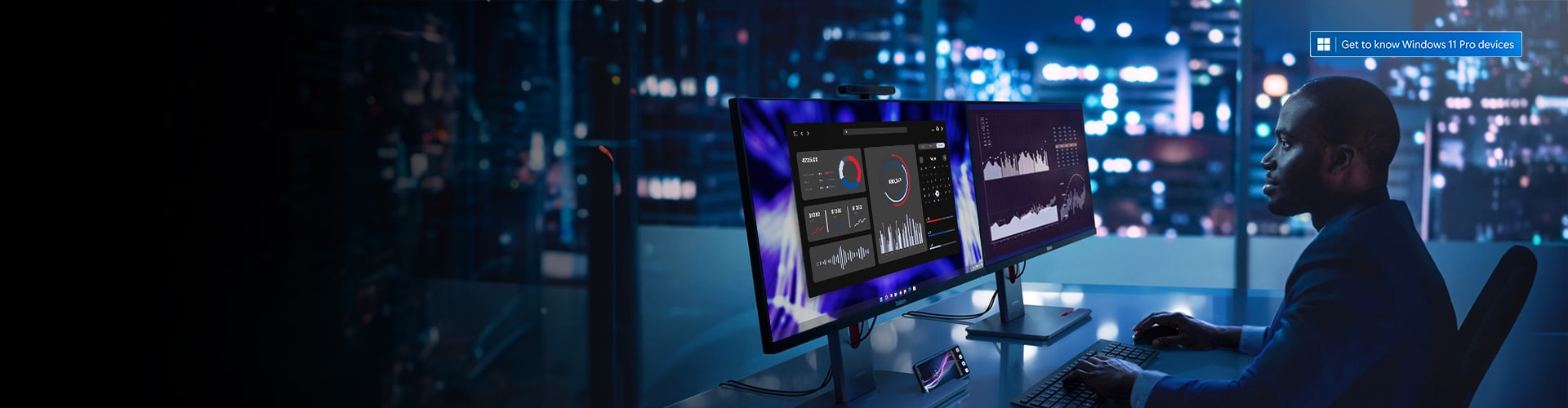 A modern office setup featuring two large Lenovo ThinkCentre PCs Monitors displaying data visualizations and charts. The background shows a cityscape at night through large windows.
