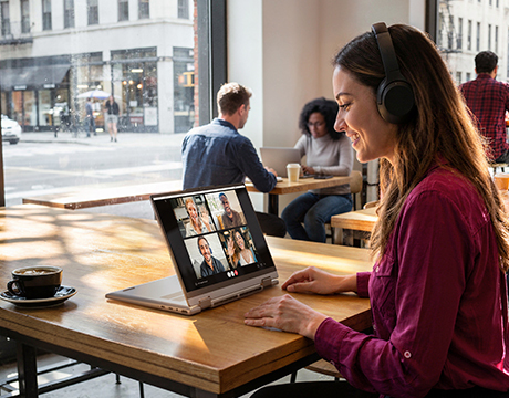 Woman with headphones video conferencing in a cafe, demonstrating the portability of the Lenovo ThinkBook 14 2-in-1 Gen 6 Laptop.
