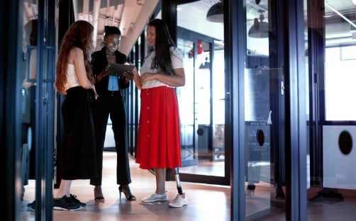 Three women standing in a hallway looking at a tablet that the lady in the back is holding.