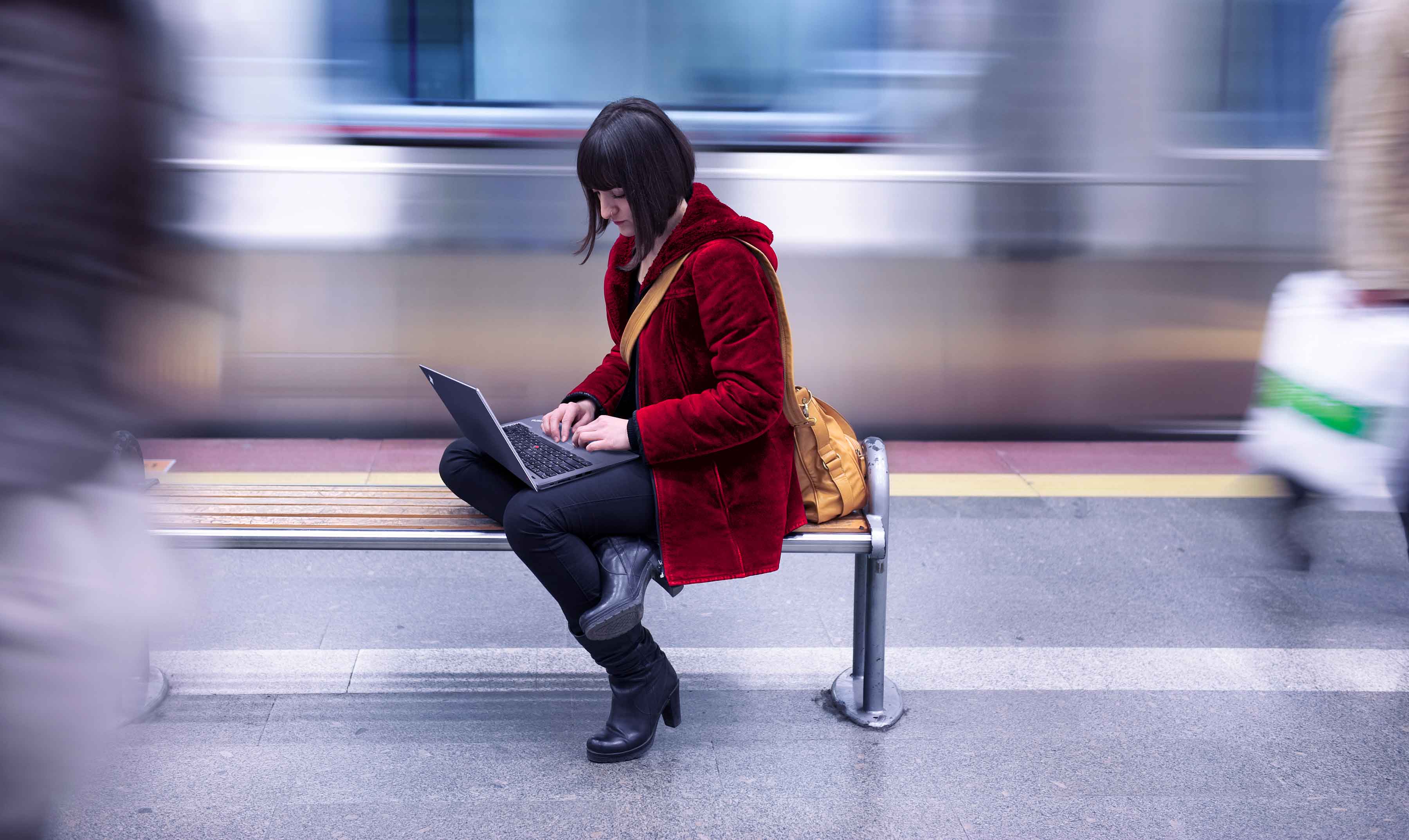 A woman working on a Lenovo laptop while sitting on a bench in a subway station, as a train zooms past behind her.