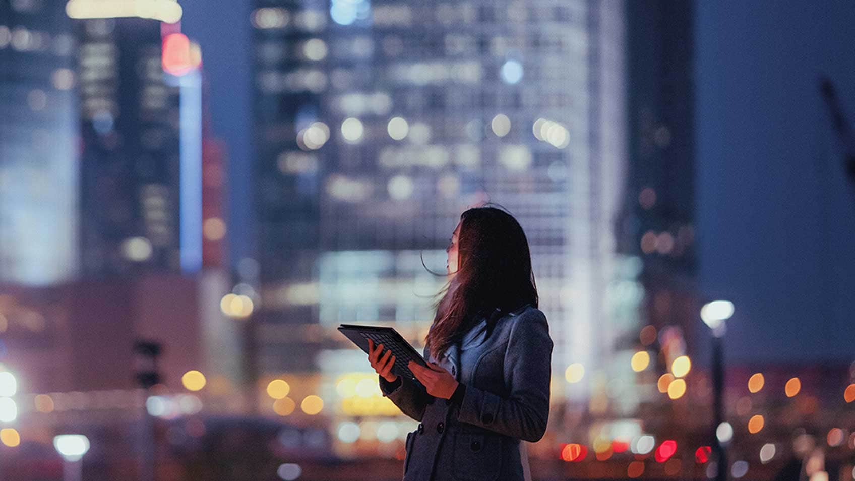 Une femme tenant une tablette et regardant par-dessus son épaule la ville derrière elle.