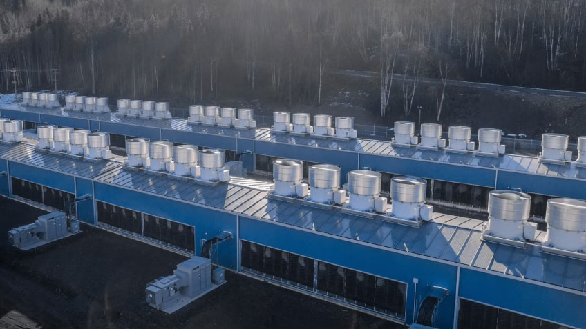 Aerial view of a blue data center with silver roof vents and transformers, set against a cold forest.