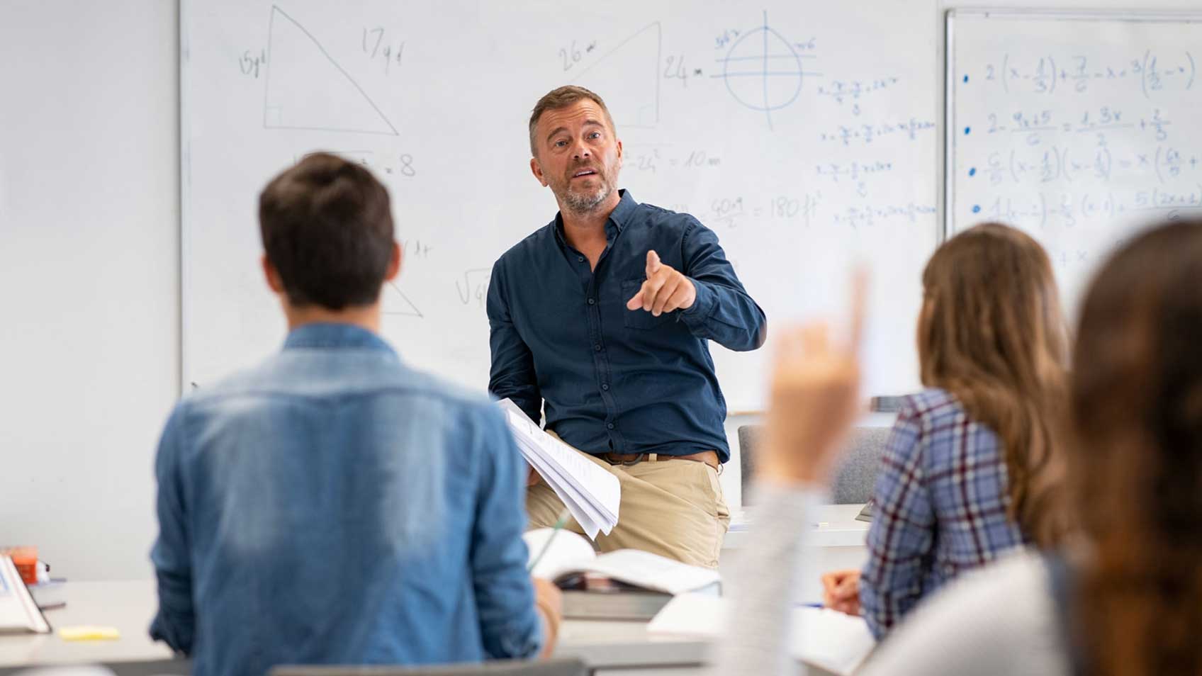 Un enseignant, devant la classe, convoquant un élève qui a la main en lève. 