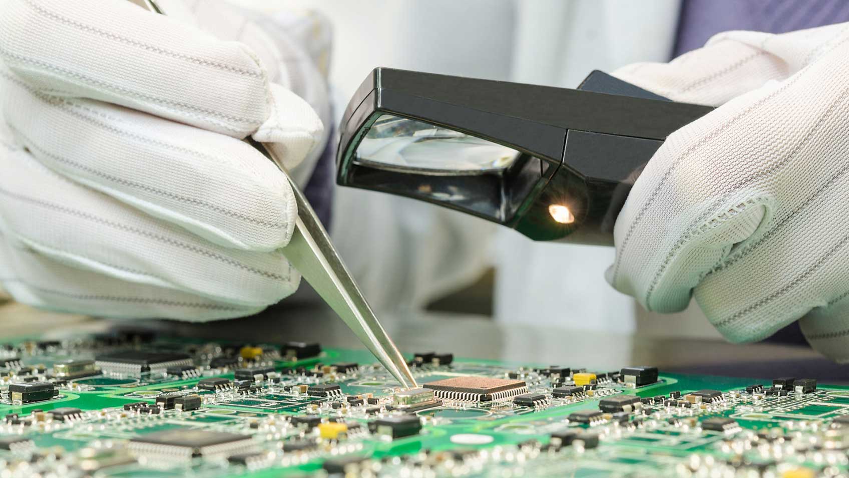 Mains gantées dans une lentille loupe et avec une pince à épiliser sur une carte de circuit imprimé.
