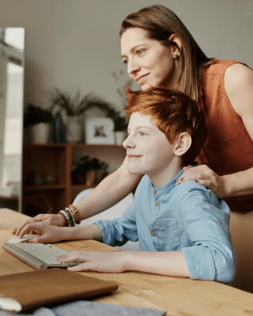 A woman rests her hand on a child’s shoulder as they both smile while looking at a computer display.