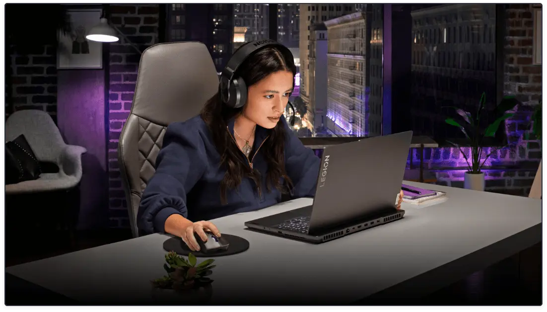 In an urban apartment with purple brick walls, a seated figure in headphones gazes intently at a Lenovo Legion gaming laptop.