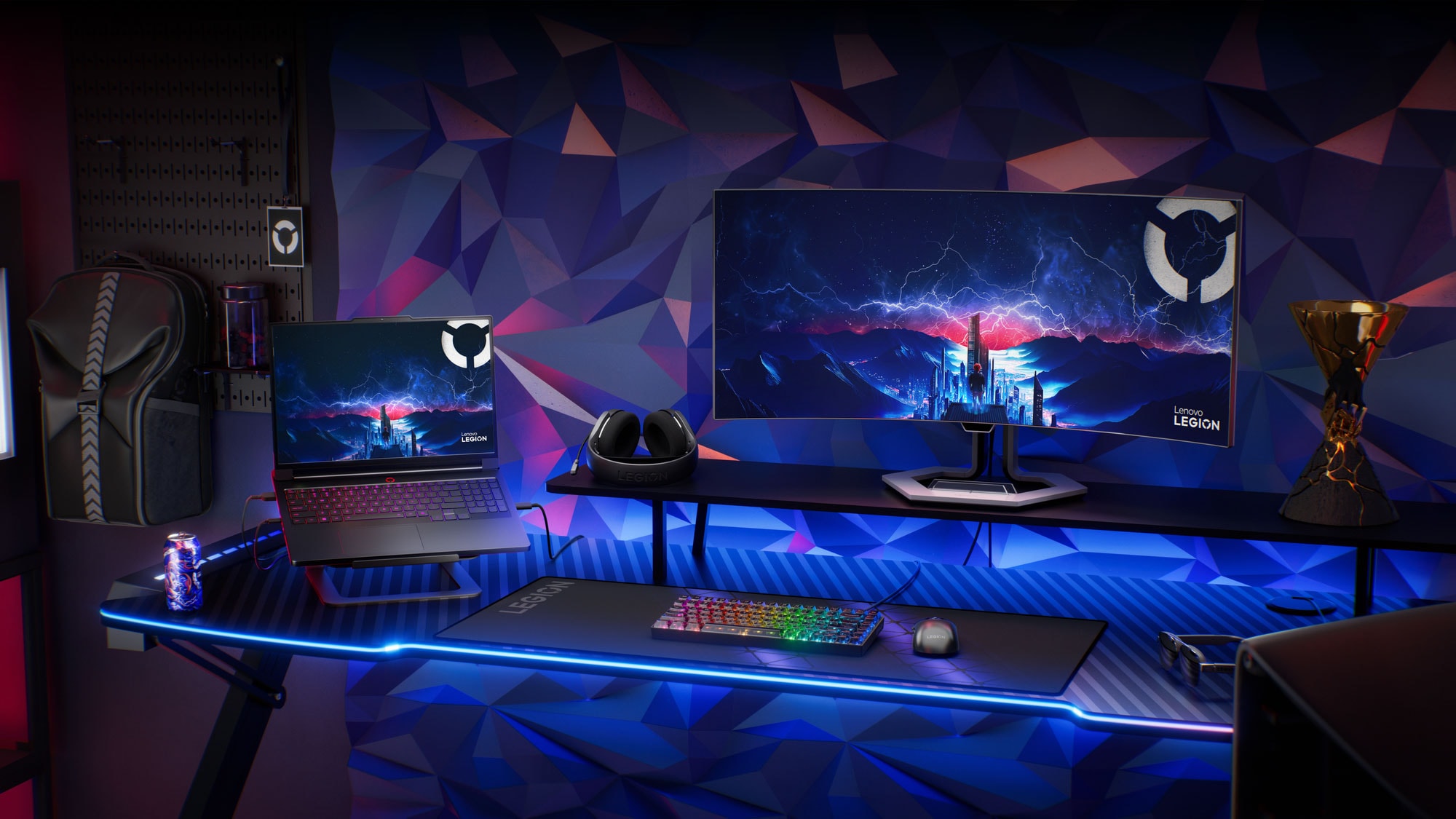 Configuration de l’écosystème de jeux Lenovo Legion comprenant un portable, un appareil portable, un moniteur incurvé, un casque, une souris, un clavier et des accessoires sur un bureau éclairé à deux tons rouge et bleu.