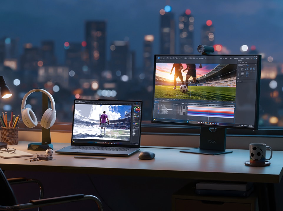 Un ordinateur Yoga, un écran, une souris et un casque reposent sur un bureau face à la ville, près d'une tasse et d'un porte-clés football. Les écrans de l'ordinateur portable et du moniteur affichent des joueurs de football sur le terrain, dans des stades.